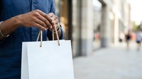 Man holding white shopping bag on urban street with blurred storefront background, retail and consumer lifestyle concept for commerce and retail design.