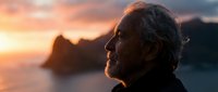 Senior man with gray beard contemplating sunset over ocean mountains. Peaceful retirement lifestyle concept for wellness and mindfulness content.