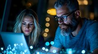 Mature bearded man and blonde woman working late on laptop with glowing screen, bokeh lights in background. Business team collaboration at night.
