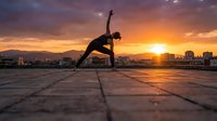 Young athletic woman practicing yoga triangle pose on urban rooftop during golden hour sunset with city skyline backdrop for wellness and fitness lifestyle.