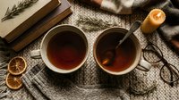 Two cozy mugs of hot tea on a knitted blanket with dried oranges, rosemary, candle, books, and glasses in warm autumn style.