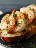 Garlic butter shrimp served on crusty bread, garnished with fresh herbs and red pepper flakes. Close-up view shows juicy seafood with delicate seasoning.