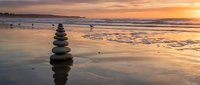 Balanced stone cairn on wet beach sand reflecting golden sunset sky over calm ocean waves creating peaceful zen meditation atmosphere for wellness concepts.