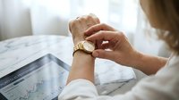 Professional businesswoman checking luxury gold wristwatch while reviewing financial charts and market data on tablet during morning work routine.