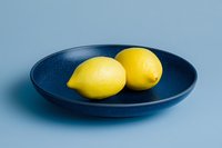 Two fresh yellow lemons on a dark blue ceramic plate against a light blue background, minimalist food styling for culinary and lifestyle projects.