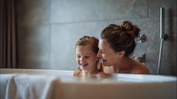 Mother and young daughter enjoying bath time together in modern bathroom, bonding moment between parent and child in warm family setting.