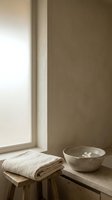 Minimalist spa bathroom corner with folded cream linen towel on wooden stool, concrete bowl with floating flower, and natural light from window for wellness and relaxation design.