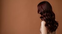 Woman with long, voluminous wavy burgundy hair styled in loose waves, shown in profile against warm brown background. Suitable for hair care, beauty, and salon marketing.