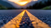Yellow road marking lines on asphalt highway at dramatic sunset with golden light reflection, close-up view with shallow depth of field, empty rural road.