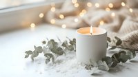 White ceramic candle burning surrounded by eucalyptus leaves on soft textured surface with warm bokeh lights creating cozy hygge atmosphere for relaxation.