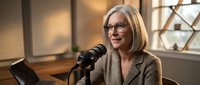 Professional middle-aged Caucasian businesswoman with blonde bob haircut and glasses speaking into podcast microphone in modern office studio setting.