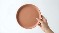 Hand holding a round terracotta ceramic plate against a white background, showcasing minimalist tableware design for food styling and home decor.