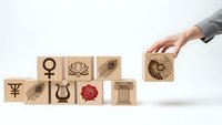Hand placing wooden cube with sun symbol among blocks featuring goddess Venus rose lyre column and lotus icons on white background for branding design.