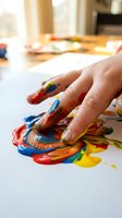 Child hand finger painting with colorful acrylic paints on white paper creating abstract art in bright classroom setting for creative education activities.