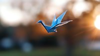 Blue origami paper crane in flight against blurred natural background with warm sunset light and bokeh effect creating magical atmosphere.