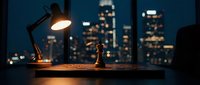 Desk lamp illuminating workspace at night with cityscape skyline visible through window, office environment for work and productivity.