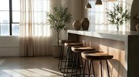 Modern kitchen counter with wooden bar stools, pendant lights, and potted plants in bright natural light from windows with sheer curtains.