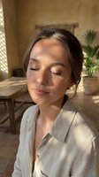 Young woman with dark hair and light skin tone wearing a light linen shirt, eyes closed in serene expression, indoor spa or wellness setting with potted plants and natural sunlight for relaxation and beauty content.