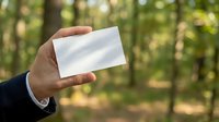 Hand of a businessman in a suit holding a blank white card outdoors in a green forest, mockup for branding or text.