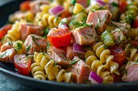 Fresh rotini pasta salad with chunks of salmon, cherry tomatoes, red onion and dill garnish served on dark plate, close up view of colorful Mediterranean dish.