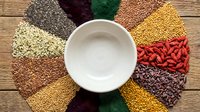 Overhead view of white ceramic bowl surrounded by colorful dried legumes and grains including lentils, beans, and seeds arranged on wooden surface for food styling and culinary content.