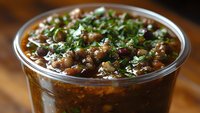 Hearty black bean soup garnished with fresh cilantro and spices in clear plastic container, rich brown broth with visible beans and vegetables, ready to serve.
