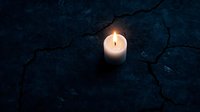 Single white candle glowing in darkness on cracked dark blue stone surface creates dramatic atmospheric mood for spiritual or memorial concepts.
