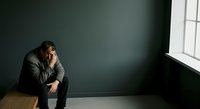 Stressed businessman in dark suit sitting alone on wooden floor against gray wall, hand on face, expressing worry, depression, or burnout in minimalist office environment.