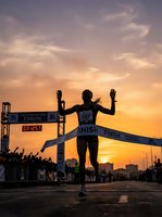 Silhouetted runner crossing finish line at sunset marathon event with golden sky and spectators lining street, celebrating athletic achievement and endurance sports competition.
