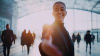 Confident young Asian businessman smiling at camera in modern transit hub with sunlight flare, surrounded by commuters during rush hour.