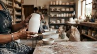 Artisan potter holding finished ceramic vessel in studio workshop with shelves of pottery supplies and works in progress, natural light from window.