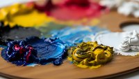 Artist palette with vibrant oil paints in blue, yellow, red and white colors mixed on wooden surface for creative painting and art projects.