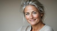 Confident mature Caucasian woman with silver gray hair and natural makeup smiling warmly at camera against neutral background, portraying wisdom and authenticity.