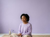 Young Black woman with shoulder-length braided hair wearing lavender sweater, sitting at wooden table with potted plant against purple wall background for wellness and lifestyle content.