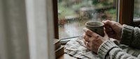 Cozy morning scene with hands holding warm coffee mug by rainy window, perfect for lifestyle and comfort themes in marketing materials.