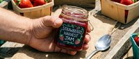 Hand holding homemade strawberry jam jar at farmers market with fresh berries in wooden baskets and spoon on rustic table for food preservation and artisan product sales.