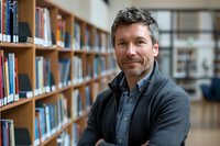 Confident middle-aged Caucasian man with gray hair smiling in library, wearing casual blazer and button-up shirt against blurred bookshelf background.