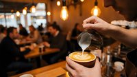 Barista pouring steamed milk into espresso cup creating latte art in warm cafe setting with blurred background of customers and hanging lights, suitable for coffee shop marketing and hospitality content.
