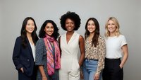 Diverse group of five professional women standing together smiling in business casual attire against neutral background for teamwork concepts.