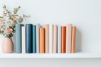 Books in pastel blue and terracotta shades arranged on white shelf with eucalyptus in ceramic vase, creating minimal contemporary home decor composition.
