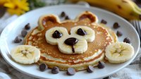 Cute bear-shaped pancake decorated with banana slices, chocolate chips creating playful animal face on white plate, fun breakfast food styling for children.