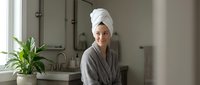 Young woman in bathrobe with towel on head relaxing in modern bathroom after shower. Spa day at home wellness routine for skincare and self-care lifestyle.