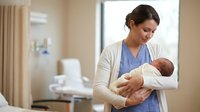 Young Hispanic female nurse in blue scrubs holding newborn baby in modern hospital maternity ward for healthcare and childcare concepts.