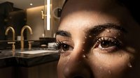 Close-up of a woman's face with dramatic eye makeup and glowing skin, looking directly at camera in modern bathroom setting with warm lighting.