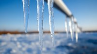 Frozen icicles hanging from roof edge against bright blue winter sky and snowy landscape background, natural crystalline ice formations catching sunlight.
