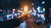 Hand holding glowing vintage Edison bulb against blurred city lights at night representing innovation, creativity and bright ideas for business concepts.