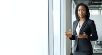 Young woman of African descent wearing professional dark blazer and white shirt, standing in modern office environment near glass windows, gesturing while speaking or presenting to colleagues.