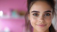 Young woman with freckles and natural makeup smiling warmly at camera against pink background, close-up portrait showcasing genuine happiness and confidence.