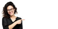 Young woman with curly hair and glasses wearing black top pointing to side with cheerful smile against white background for business or education concept.