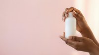 Young adult with medium brown skin tone holding white cylindrical bottle against soft pink background, suitable for cosmetics, skincare, lotion, serum, or beauty product packaging mockups.